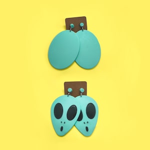 Op de afbeelding: Twee paar turquoise blauwe oorbellen. Het ene paar is rond en het andere paar is gevormd als alienhoofden met zwarte ogen.
