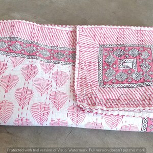 May include: A pink and white kantha quilt with a leaf pattern and a geometric design in the center. The quilt is folded in half and has a white fringe around the edges.