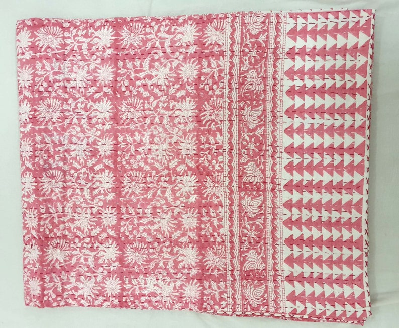 K&ouml;nnte beinhalten: Eine rosa und wei&szlig; gemusterte Steppdecke. Die Steppdecke hat auf der linken Seite ein sich wiederholendes Blumendesign, in der Mitte einen dekorativen Rand und auf der rechten Seite ein geometrisches Dreieckmuster. Die Steppdecke besteht aus einem weichen, gesteppten Stoff.