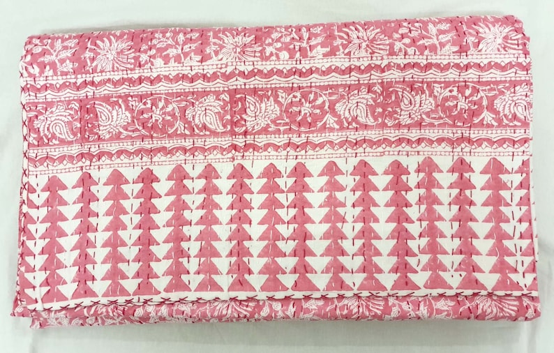 K&ouml;nnte beinhalten: Eine gefaltete rosa und wei&szlig;e Steppdecke mit einem geometrischen und floralen Muster. Die Steppdecke hat ein sich wiederholendes Dreieckdesign im unteren Bereich und Blumen- und Paisley-Muster im oberen Bereich. Die Steppdecke ist handgen&auml;ht.