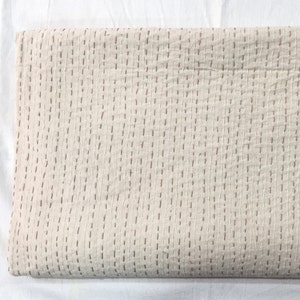 Könnte beinhalten: Eine weiße Kantha-Steppdecke mit braunen Nähten. Die Decke ist in der Hälfte gefaltet und hat eine glatte, strukturierte Oberfläche.