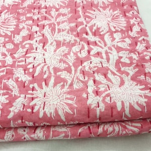 K&ouml;nnte beinhalten: Gefalteter rosa Stoff mit wei&szlig;em Blumenmuster. Gro&szlig;e wei&szlig;e Blumen und Bl&auml;tter sind auf dem rosa Hintergrund aufgedruckt. Der Stoff ist mit rotem Faden gen&auml;ht, was ihm ein gestepptes Aussehen verleiht. Thema: Heimdekoration.
