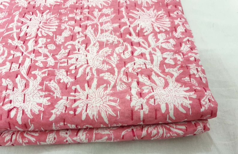 K&ouml;nnte beinhalten: Gefalteter rosa Stoff mit wei&szlig;em Blumenmuster. Gro&szlig;e wei&szlig;e Blumen und Bl&auml;tter sind auf dem rosa Hintergrund aufgedruckt. Der Stoff ist mit rotem Faden gen&auml;ht, was ihm ein gestepptes Aussehen verleiht. Thema: Heimdekoration.