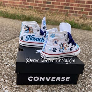May include: White Converse high-top sneakers customized with Bluey cartoon characters, the name "Nevaeh", and blue paw prints. The shoes have blue ribbon laces and are decorated with blue and clear rhinestones. The shoes are on top of a black Converse box.