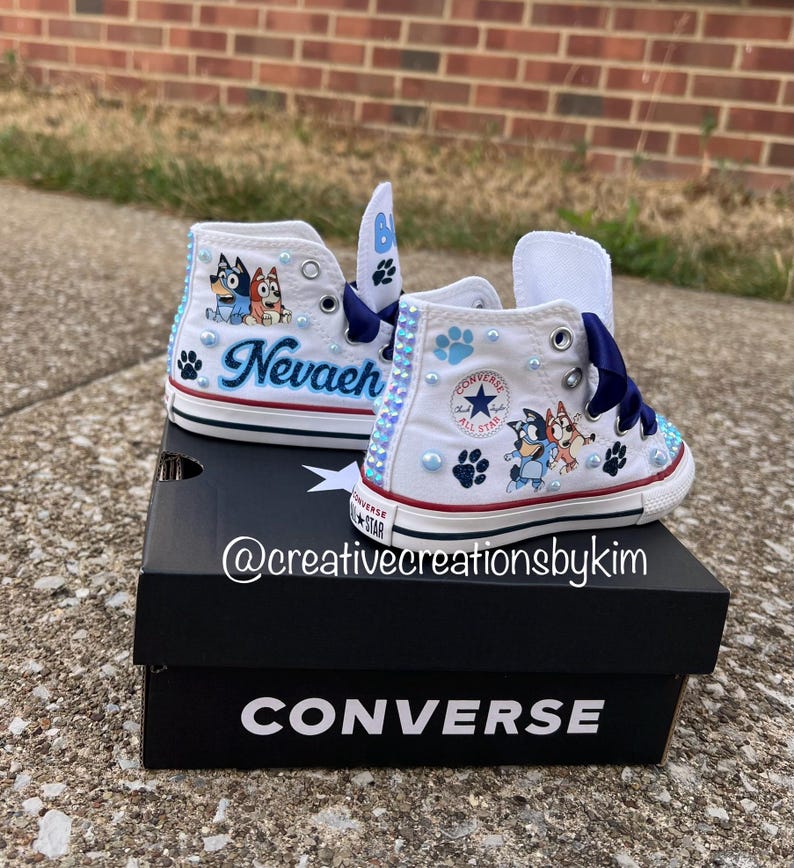 May include: White Converse high-top sneakers customized with Bluey cartoon characters, the name "Nevaeh", and blue paw prints. The shoes have blue ribbon laces and are decorated with blue and clear rhinestones. The shoes are on top of a black Converse box.