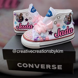 May include: White high-top Converse sneakers customized with the name "Jada" in pink script. The shoes feature images of characters from the movie Frozen, pink bows, and decorative gems. The shoes are on a black Converse box.