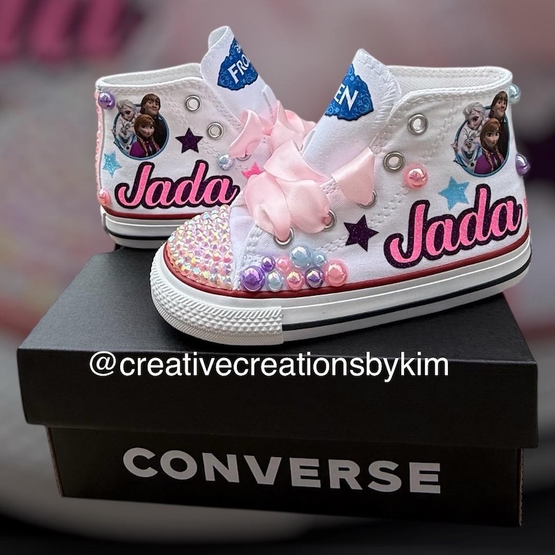 May include: White high-top Converse sneakers customized with the name "Jada" in pink script. The shoes feature images of characters from the movie Frozen, pink bows, and decorative gems. The shoes are on a black Converse box.