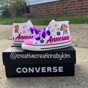 May include: White high-top sneakers with purple ribbon laces, featuring a cartoon character and the name "Annessa" in pink. The shoes are decorated with pink gems and paw prints, sitting on a black Converse box.