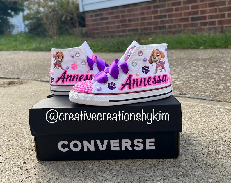 May include: White high-top sneakers with purple ribbon laces, featuring a cartoon character and the name "Annessa" in pink. The shoes are decorated with pink gems and paw prints, sitting on a black Converse box.