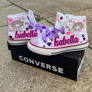 May include: White high-top Converse sneakers customized with the name "Isabella" in pink script. The shoes feature rainbow and heart designs, purple ribbon laces, and decorative embellishments. They sit on a black Converse box.