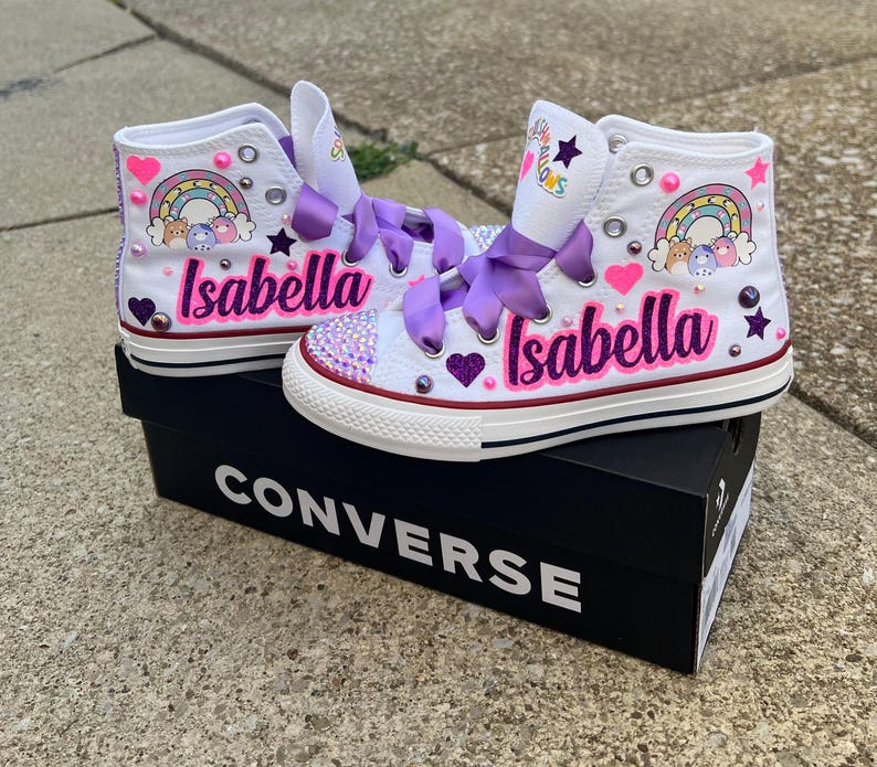 May include: White high-top Converse sneakers customized with the name "Isabella" in pink script. The shoes feature rainbow and heart designs, purple ribbon laces, and decorative embellishments. They sit on a black Converse box.