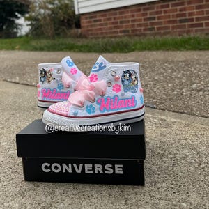 May include: White high-top sneakers with custom designs, including pink bows, paw prints, and the name "Milani" in pink and blue. The toe caps are embellished with pink sequins. The shoes are displayed on a black Converse box.