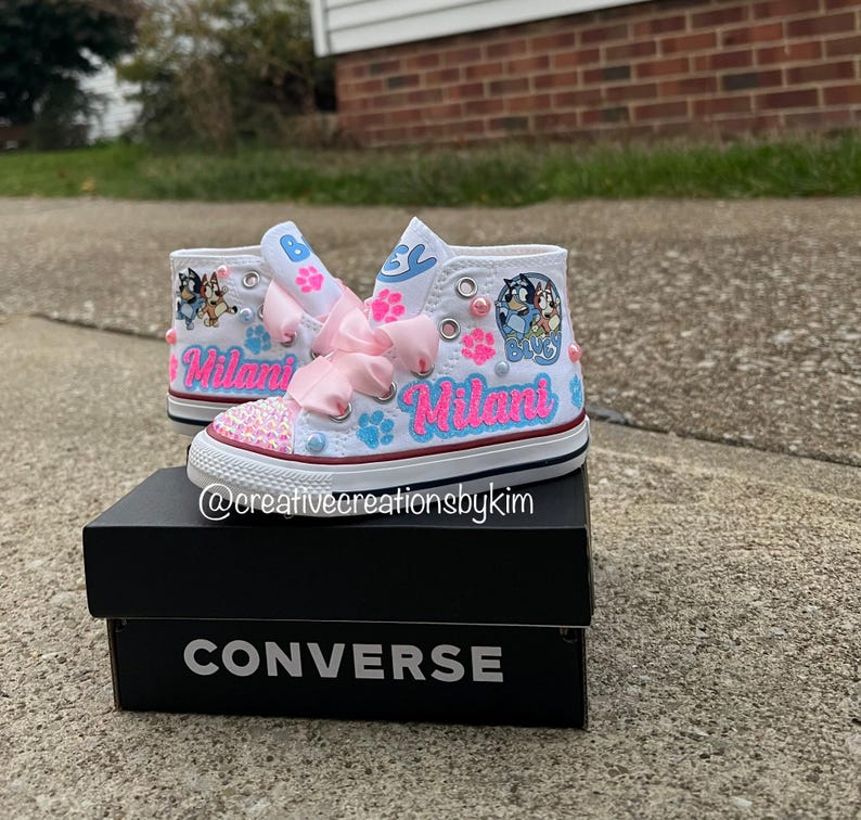 May include: White high-top sneakers with custom designs, including pink bows, paw prints, and the name "Milani" in pink and blue. The toe caps are embellished with pink sequins. The shoes are displayed on a black Converse box.