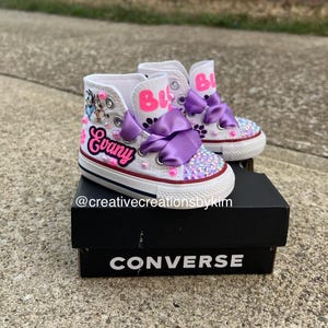 May include: White high-top sneakers with custom designs. The shoes feature the name "Evany" in pink script, purple bows, and sparkling embellishments on the toes. The shoe's tongue has the name "Bi" in pink. The shoes are on a black Converse box.