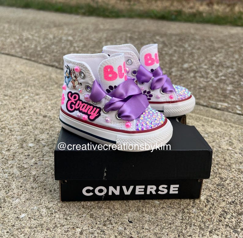 May include: White high-top sneakers with custom designs. The shoes feature the name "Evany" in pink script, purple bows, and sparkling embellishments on the toes. The shoe's tongue has the name "Bi" in pink. The shoes are on a black Converse box.