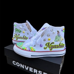 May include: White high-top sneakers with custom embellishments. The sneakers feature the name "Nyashia" in purple and green, along with images of a frog and a princess. The shoes are decorated with colorful beads and pearls, and sit on a black Converse box.