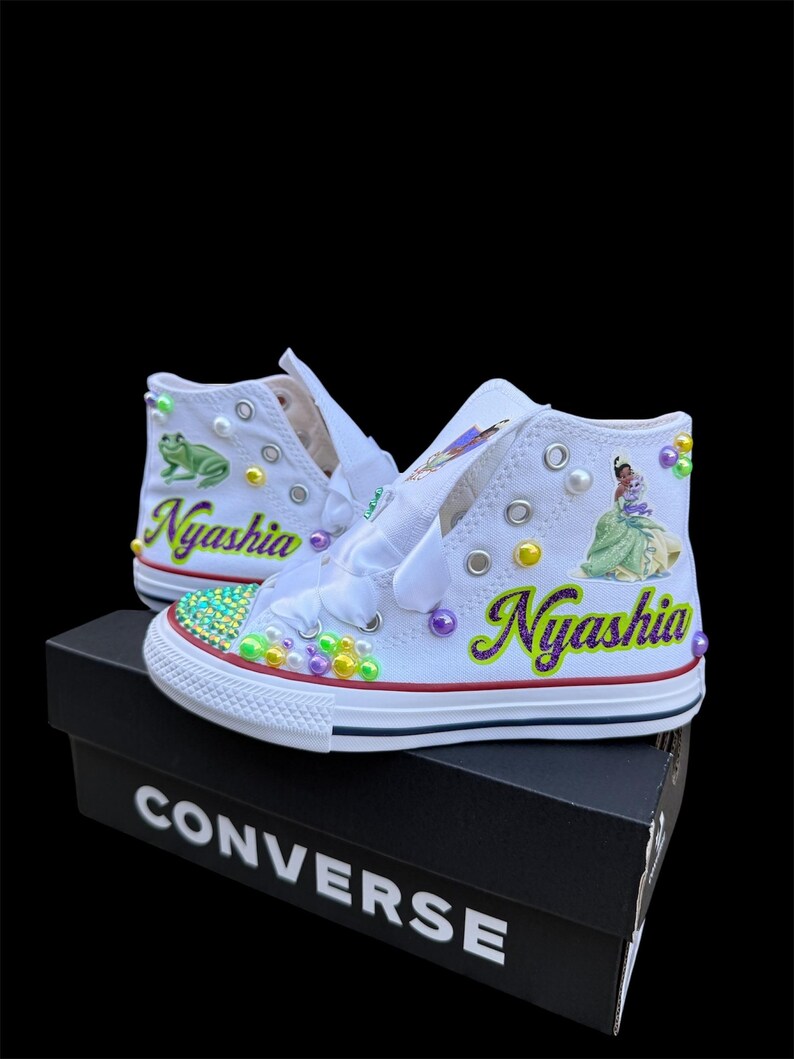 May include: White high-top sneakers with custom embellishments. The sneakers feature the name "Nyashia" in purple and green, along with images of a frog and a princess. The shoes are decorated with colorful beads and pearls, and sit on a black Converse box.