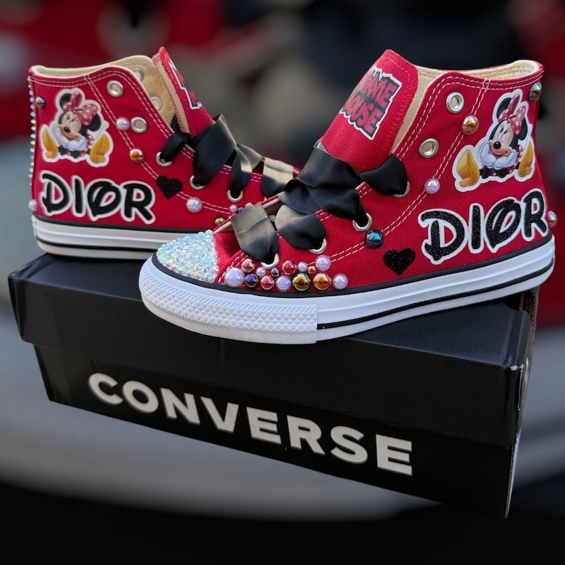 May include: Red high-top sneakers with black bows, embellished with colorful gems and Minnie Mouse graphics. The word "DIOR" is printed on the side. The sneakers are on a black Converse box.
