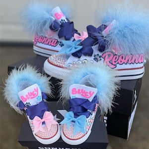 May include: White canvas sneakers with custom embellishments. The shoes feature light blue feather trim, bows, and rhinestones. One pair has the name "Reonna" in pink script, and the other pair has the name "Bluey".