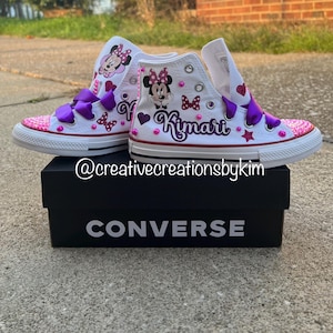 May include: White high-top Converse sneakers customized with a Minnie Mouse theme. The sneakers feature purple bows, pink sequin toes, and the name "Kimari" in purple script. The shoes are displayed on a black Converse box.