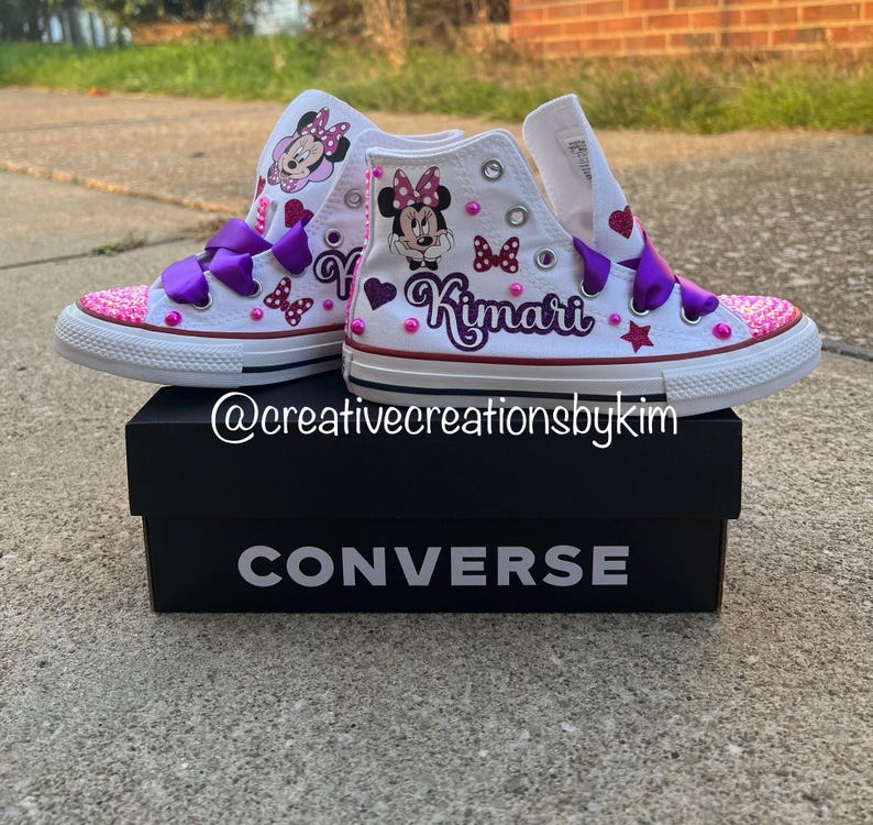 May include: White high-top Converse sneakers customized with a Minnie Mouse theme. The sneakers feature purple bows, pink sequin toes, and the name "Kimari" in purple script. The shoes are displayed on a black Converse box.