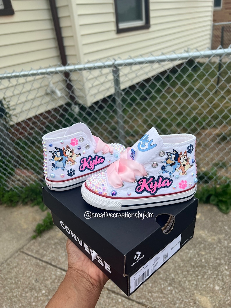 May include: White high-top sneakers customized with Bluey and Bingo cartoon characters, pink ribbon laces, and the name "Kyla" in pink script. The shoes are embellished with rhinestones and studs.