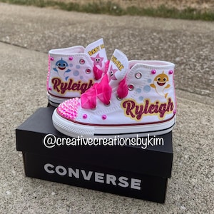 May include: White high-top sneakers with pink accents, featuring a Baby Shark theme. The shoes have pink glitter on the toes, pink laces, and the name "Ryleigh" in glitter. The shoes are on a black Converse box.
