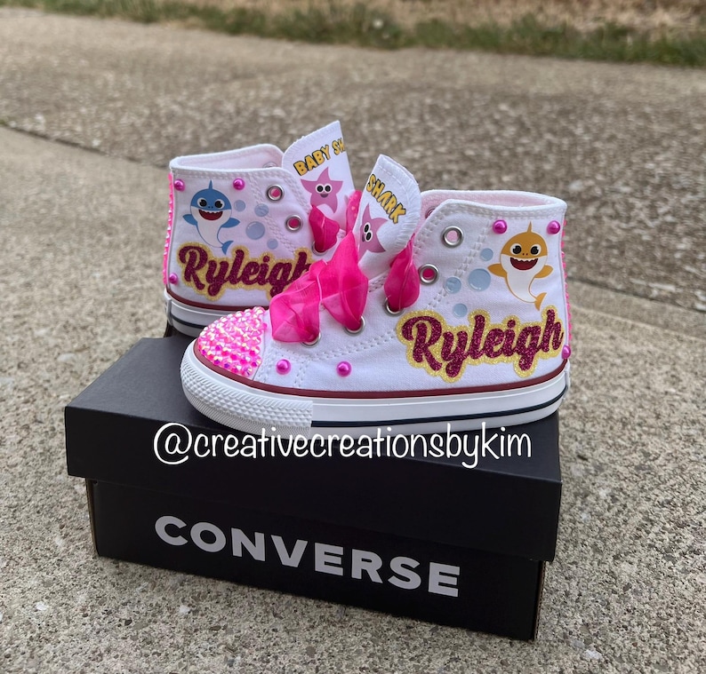 May include: White high-top sneakers with pink accents, featuring a Baby Shark theme. The shoes have pink glitter on the toes, pink laces, and the name "Ryleigh" in glitter. The shoes are on a black Converse box.