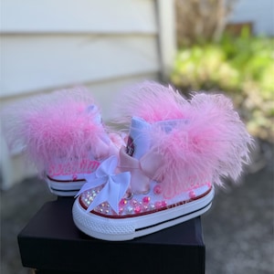 May include: White Converse sneakers with pink feather trim, embellished with pink and iridescent rhinestones and a white bow. The sneakers sit on a black Converse box.