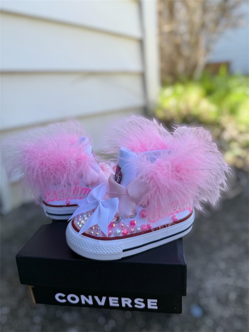 May include: White Converse sneakers with pink feather trim, embellished with pink and iridescent rhinestones and a white bow. The sneakers sit on a black Converse box.