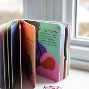 May include: A colorful children's book with the letter M and the words "M is for midwife." The illustration shows a midwife helping a person give birth. The book is open to reveal other pages with different letters and illustrations.