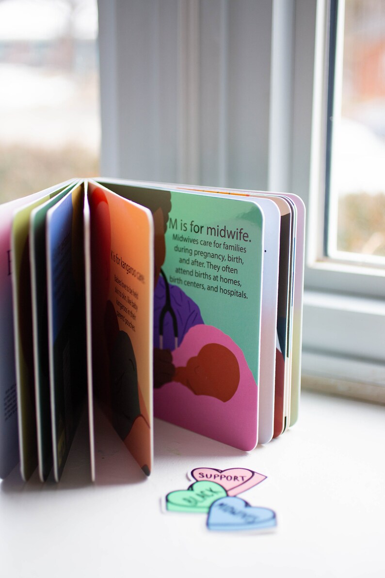 May include: A colorful children's book with the letter M and the words "M is for midwife." The illustration shows a midwife helping a person give birth. The book is open to reveal other pages with different letters and illustrations.