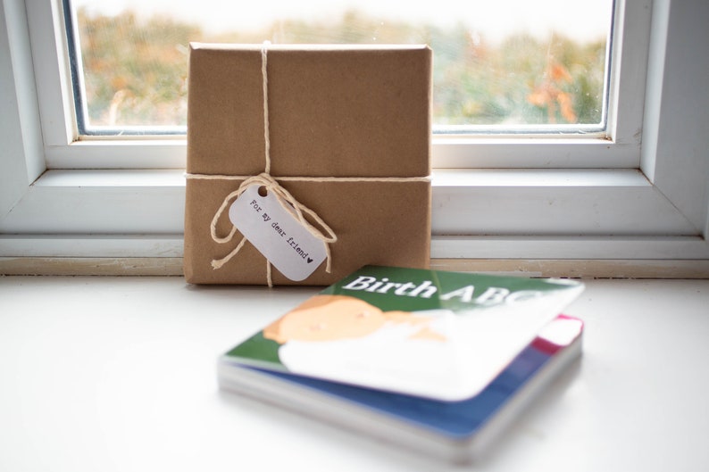 May include: A brown gift box wrapped in brown paper with twine and a white tag that says "For my dear friend" with a heart. The box is sitting on a white surface next to a green and blue book with the title "Birth ABC".