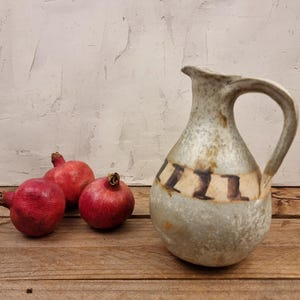 May include: A handcrafted ceramic jug with a wide handle and a decorative band of brown and cream rectangles. Three ripe pomegranates with deep red skin are arranged on a wooden surface, complementing the jug's earthy tones.