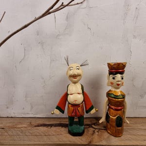 May include: Two wooden hand puppets, one with a red and orange outfit and the other with a gold and green outfit. The puppets have painted faces and are standing on a wooden surface.