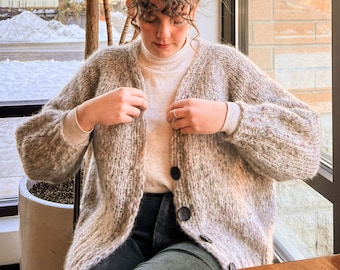 May include: A woman wearing a light gray cardigan with a white turtleneck sweater underneath. The cardigan has a speckled pattern and is knitted with a loose stitch. The woman is wearing dark green pants.