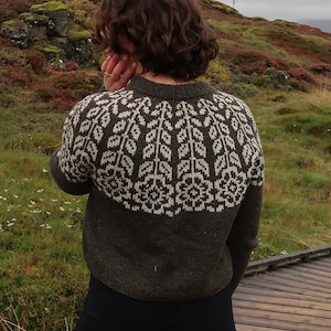 Op de afbeelding: Een donkergrijze trui met een wit bloemmotief over de schouders en de bovenrug. De trui wordt gedragen met een zwarte broek. De achtergrond toont een natuurlijk landschap met groene en bruine vegetatie.