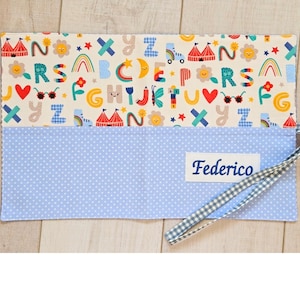 Può includere: Un oggetto rettangolare in tessuto con un design stravagante. La sezione superiore presenta uno sfondo color crema con lettere colorate, tendoni da circo, arcobaleni e altre illustrazioni giocose. La sezione inferiore è azzurra con pois bianchi e il nome "Federico" è stampato.