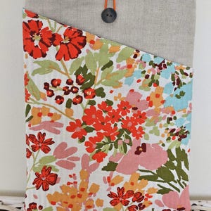 Op de afbeelding: Een tablet hoes met een bloemenpatroon en een grijze linnen bovenkant. De stof toont rode, roze, oranje en groene bloemen op een witte achtergrond. Een grijze knoop en een oranje lus sluiten de hoes af.