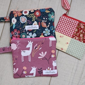 Op de afbeelding: Drie kleine stoffen zakjes met ritsen. Eén heeft een donkerblauw bloemenpatroon, een andere een roze hert- en bloemenpatroon, en de derde een patchwork-ontwerp met rood vichy, bloemen en stippen.