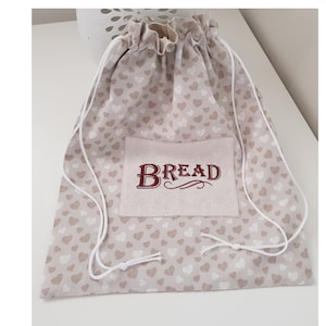 Puede incluir: Una bolsa de cordón beige con un patrón de corazones blancos. La bolsa tiene un bolsillo con la palabra "BREAD" bordada en rojo.