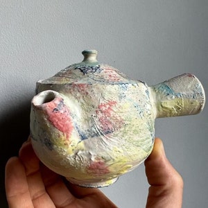 May include: A small, white ceramic teapot with a colorful, abstract design. The teapot has a rounded body, a short spout, and a handle.