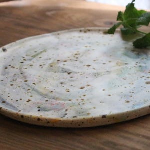 Peut inclure: Une assiette ovale en céramique, tachetée, avec une base blanc crème et de subtils motifs aquarelle bleus, verts et roses. L'assiette a un bord légèrement surélevé et est posée sur une surface en bois.