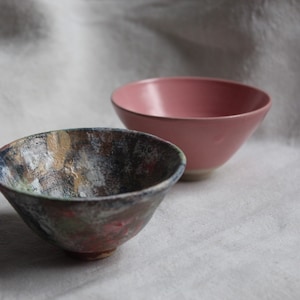 Peut inclure: Deux bols en céramique. L'un est de couleur rose uni, et l'autre est un bol texturé avec un mélange de couleurs dont le bleu, le marron et le rouge. Les bols sont posés sur un fond blanc.
