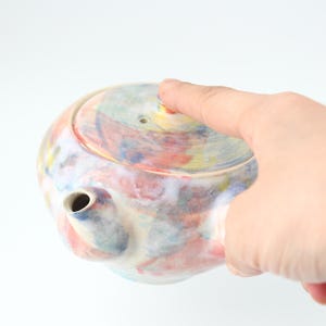 Puede incluir: Una tetera de cerámica colorida con un diseño pintado a mano en tonos de azul, rosa, amarillo y blanco. La tetera tiene un cuerpo redondo y un pico en el lateral. La tapa está ligeramente entreabierta.