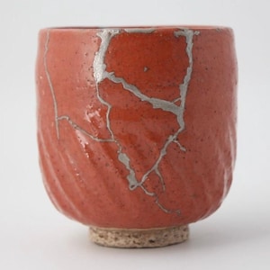 Pode incluir: Uma chávena de cerâmica vermelha com um esmalte rachado e detalhes prateados. A chávena tem uma forma arredondada e um pequeno pé.