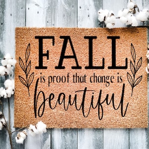 May include: A brown coir doormat with the text "Fall is proof that change is beautiful" in black lettering. The text is surrounded by two stylized leaf sprigs.
