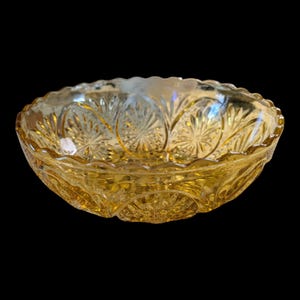 May include: A vintage amber-colored glass bowl with a scalloped rim. The bowl features a detailed pattern of starburst and leaf designs. The glass has a glossy finish, reflecting light and creating a warm, inviting aesthetic. The bowl is likely used for serving.