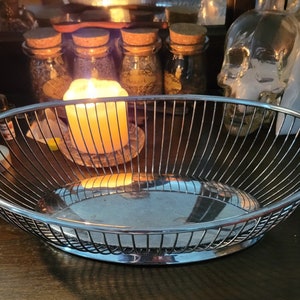 May include: A silver wire fruit bowl with a shiny, reflective surface. The bowl is empty and sits on a dark wooden surface.