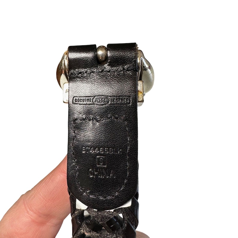 May include: Close-up of a black leather belt with a silver buckle. The belt is marked "GENUINE FOSSIL LEATHER" and "CHINA". The belt has a woven design and is held by a hand.